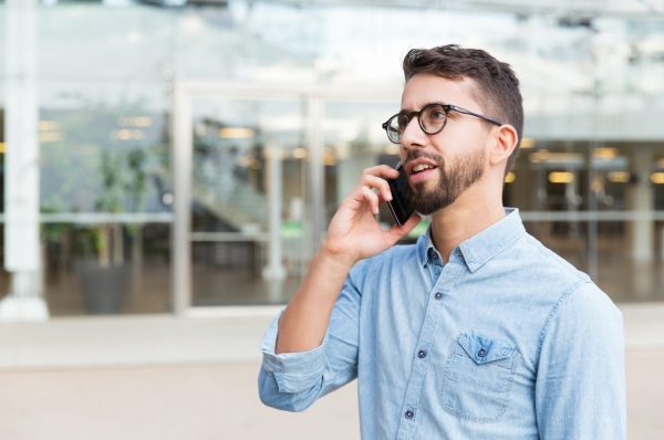 Pensive guy in eyewear speaking on cellphone and looking into distance. Handsome young man in casual shirt and glasses standing at outdoor glass wall. Phone talk concept